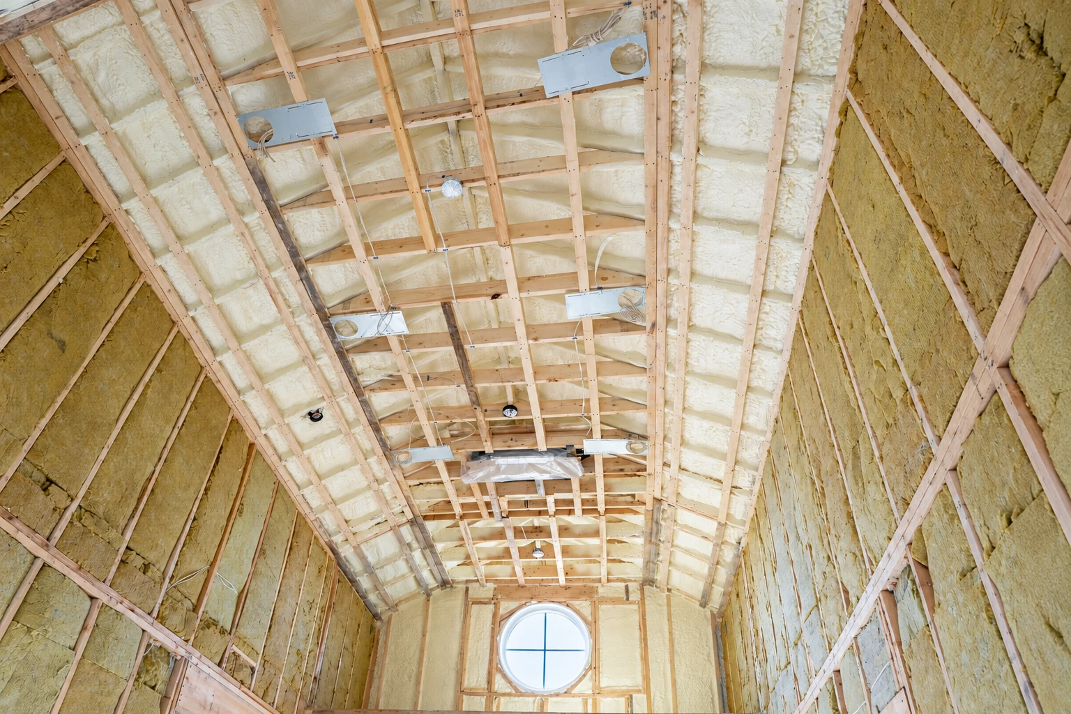 Cream-colored spray foam fills the roof and wall cavities of a tall unfinished room with exposed framing and a round window.