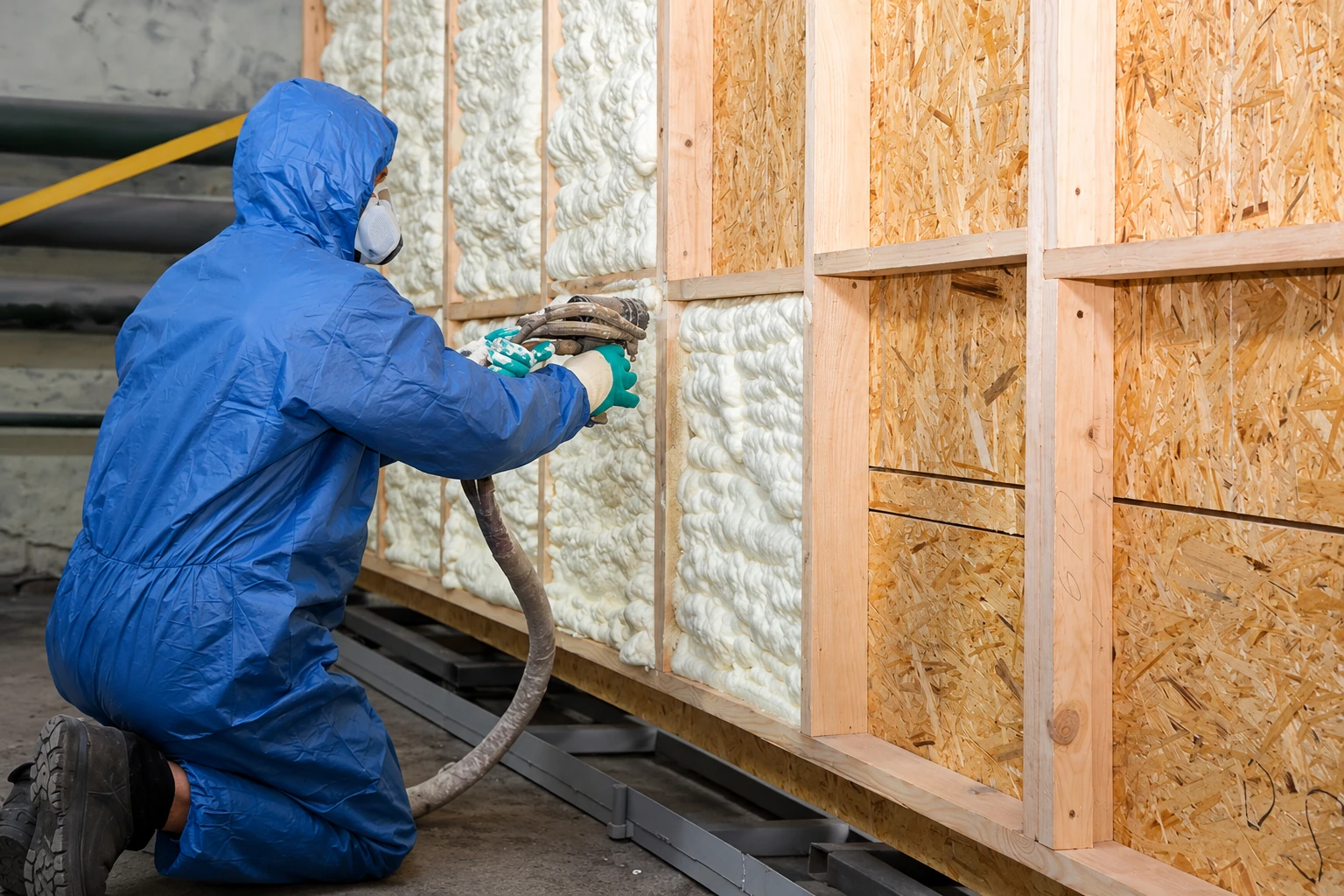 A worker in protective gear sprays foam insulation into open wall framing.