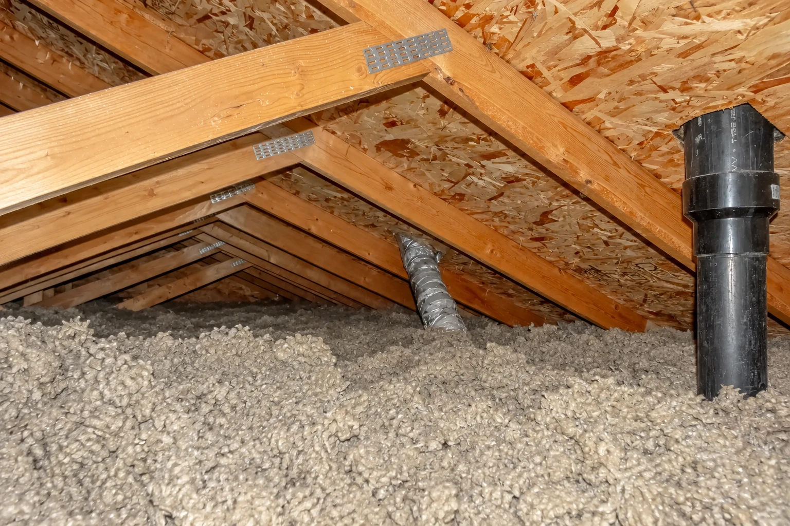 Loose-fill insulation covers an attic floor beneath exposed wooden rafters and ductwork.