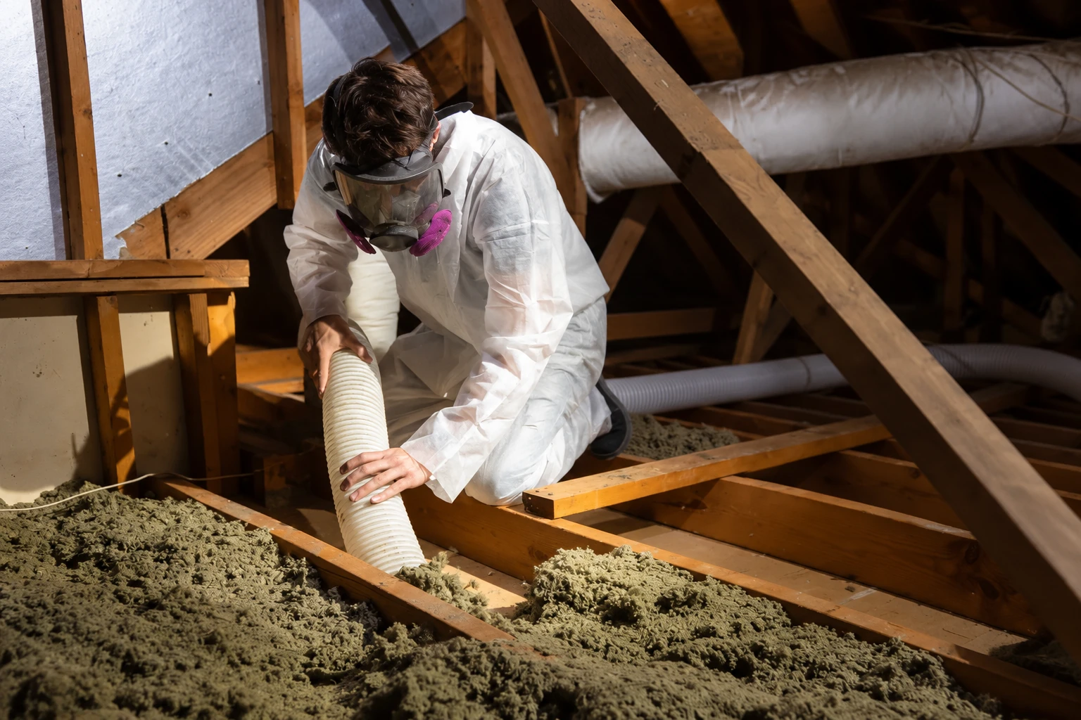 A worker in protective gear removes attic insulation using a flexible hose among exposed wooden beams.