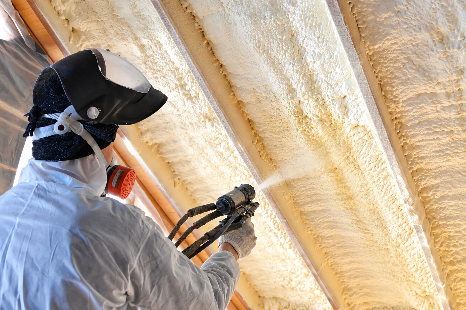 A worker in protective gear sprays foam insulation between exposed roof rafters.